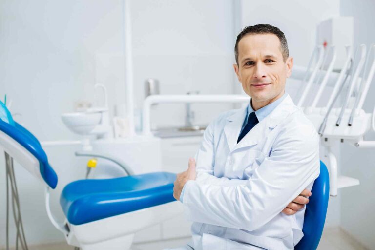 A dentist is posing for the camera in his office in white coat